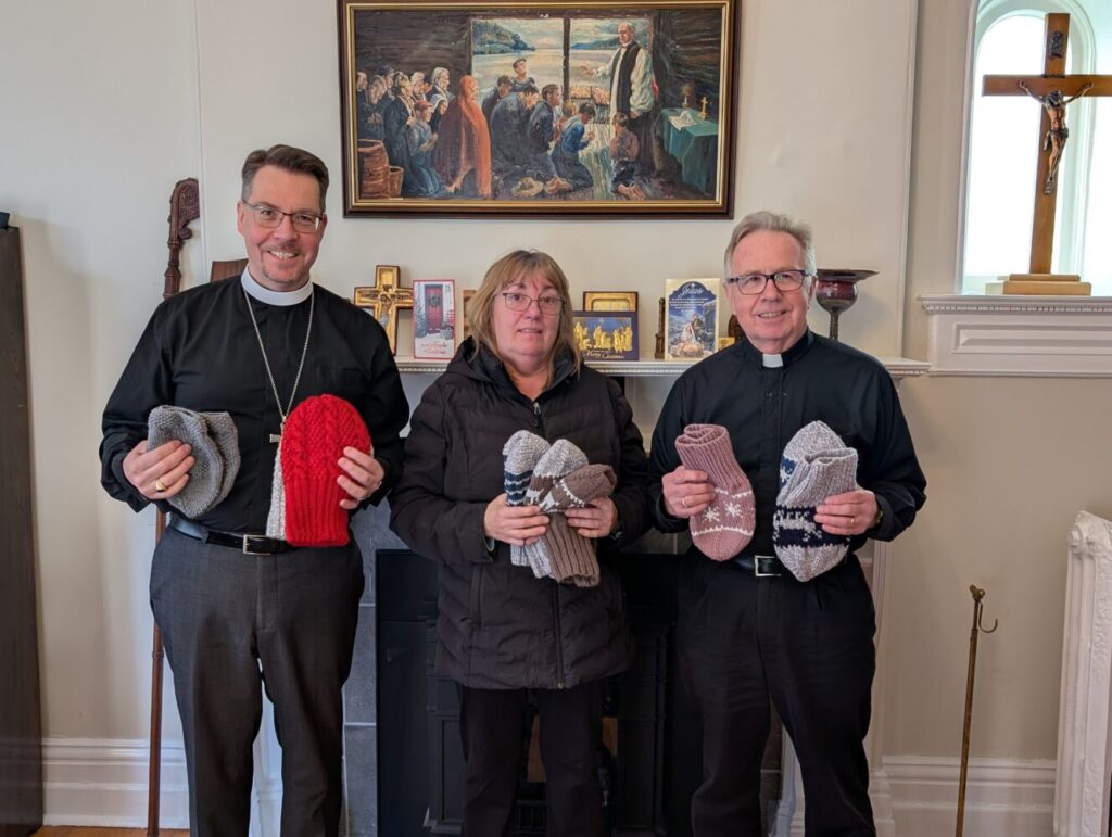 three people hold knitted goods that have been made as donations for those in need