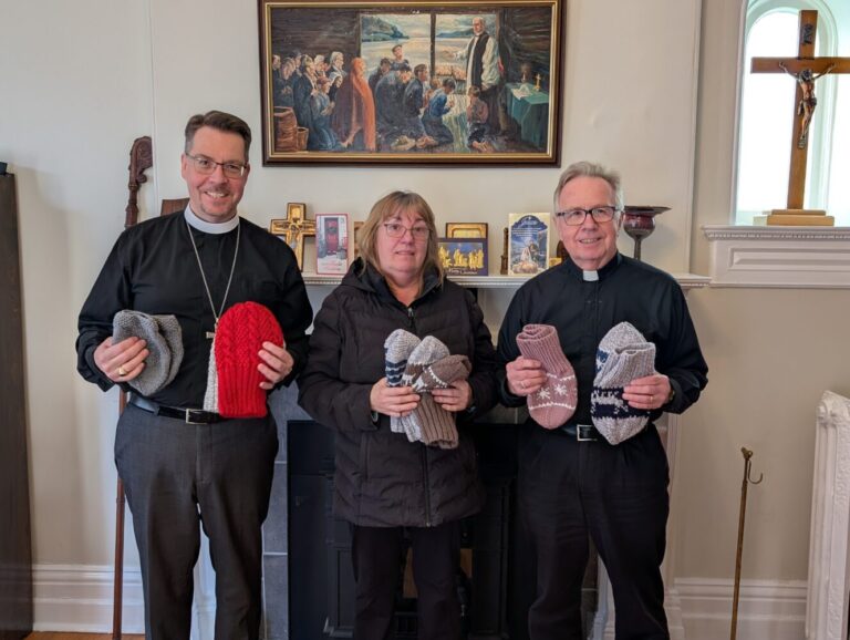 three people hold knitted goods that have been made as donations for those in need