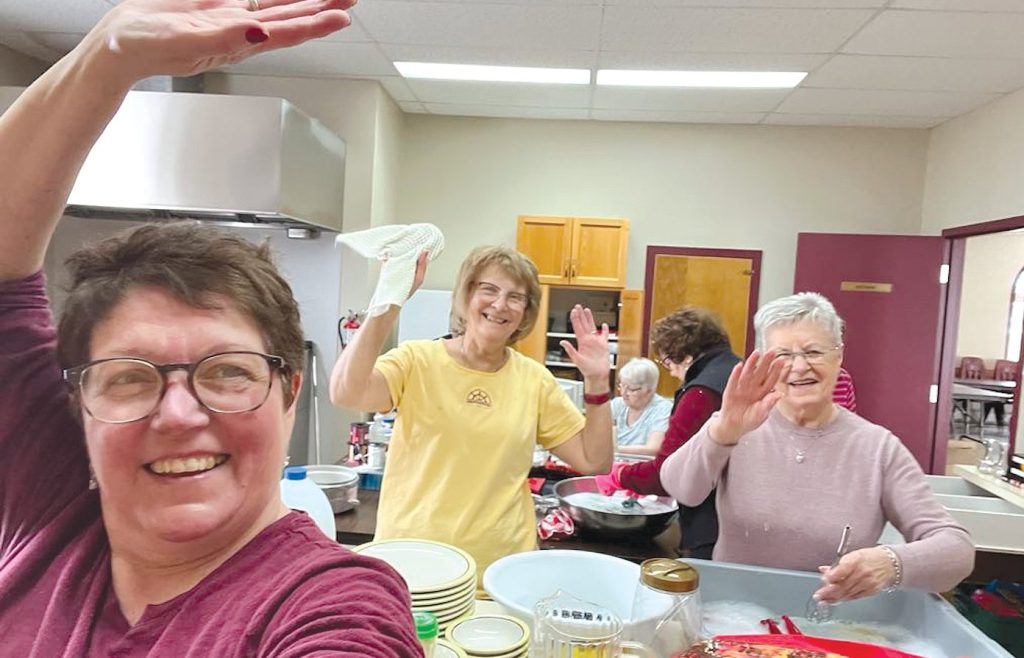 women in a kitchen at a hall, cleaning and waving at the camera