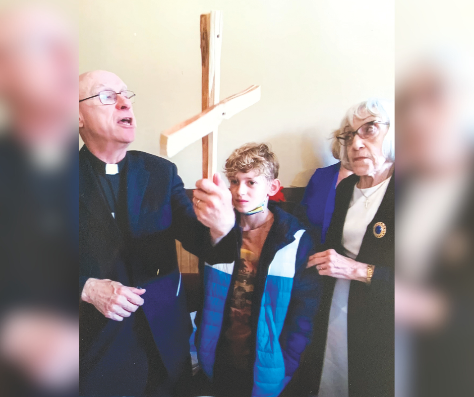 a man dressed as an Anglican priest holds a handmade cross while the young man who made it looks on. On the right is the priest's wife.