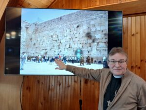 a man, dressed as a priest, points to a screen that shows an image from his trip to the Holy Land