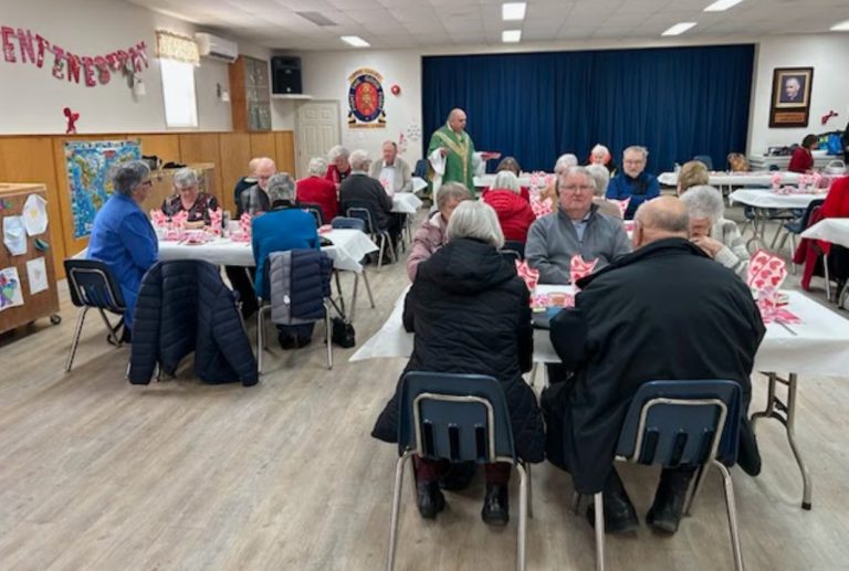 People sitting at tables in a church hall, enjoying Valentine's Day celebrations. A man dressed as a priest in green vestments in standing amongst them.