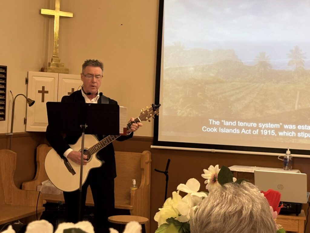 man plays the guiat in front of a screen where the words to his song appear