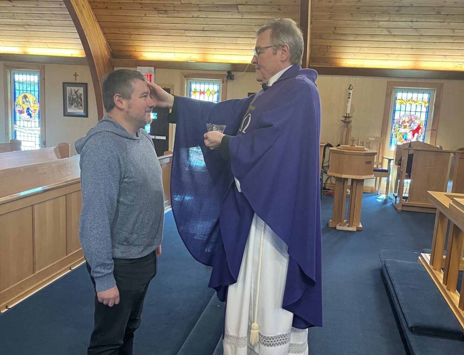 a man dressed as an Anglican priest makes the sign of the cross on another man's forehead