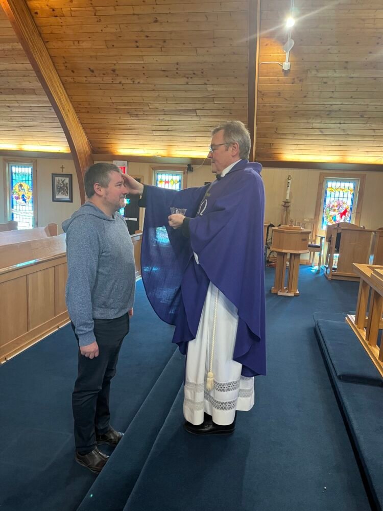 a man dressed as an Anglican priest makes the sign of the cross on another man's forehead