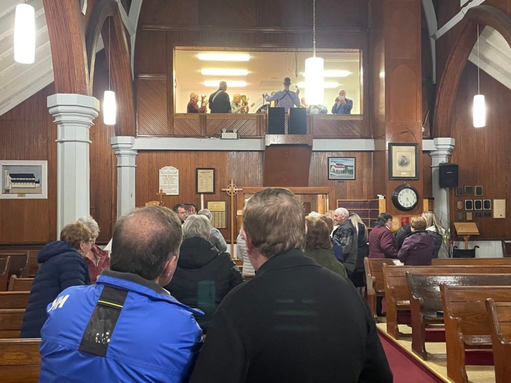many people in a church, taken from the back, who are looking up to a glassed off choir room where a band is playing.