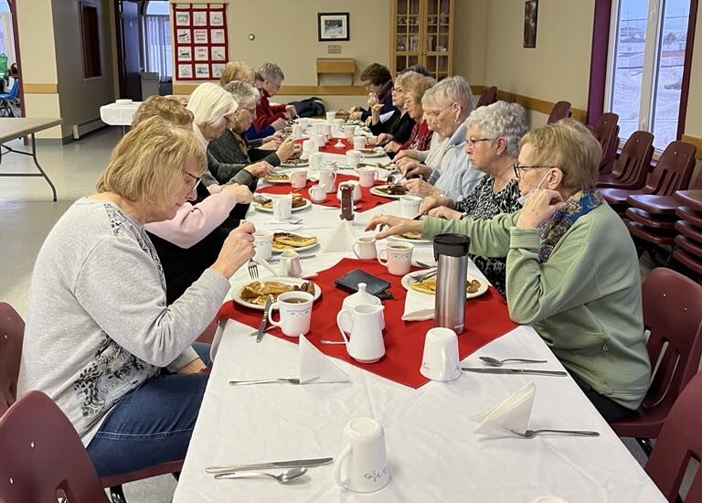 women sitting at a table in a church hall, enjoying a pancake supper