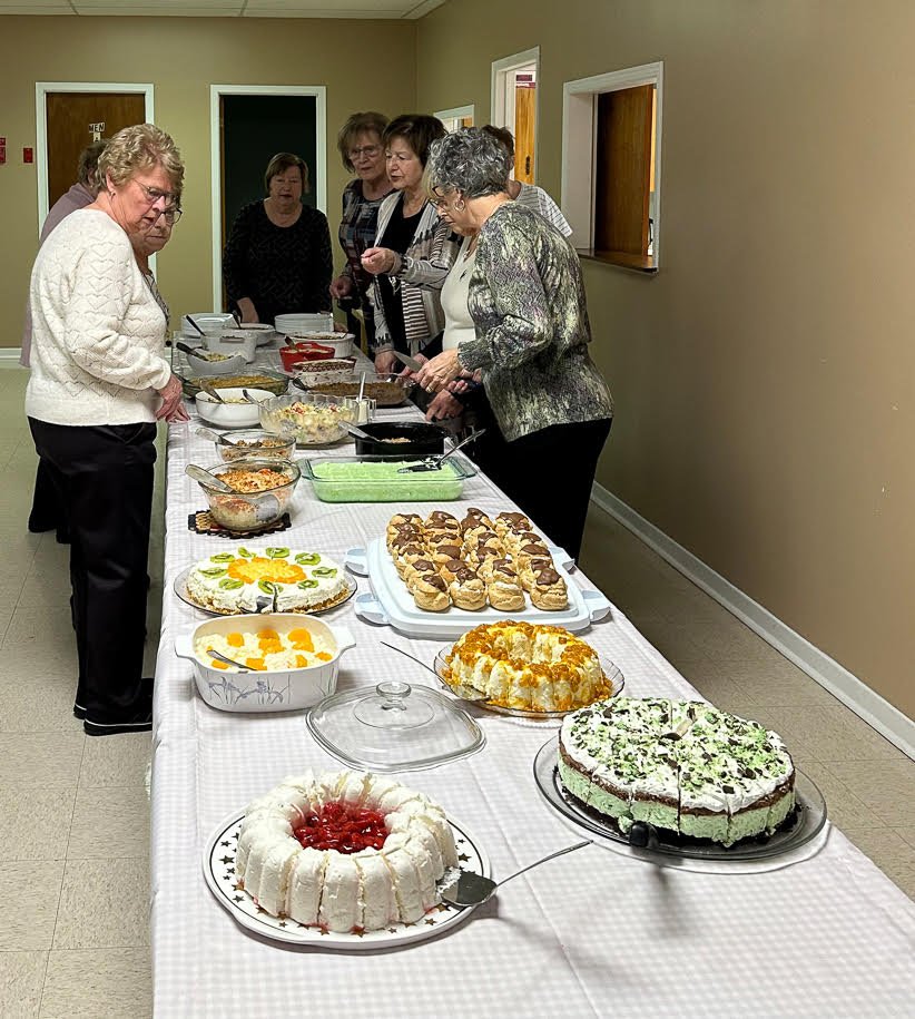People at a buffet table in a church hall with many fancy desserts on the table to chose from