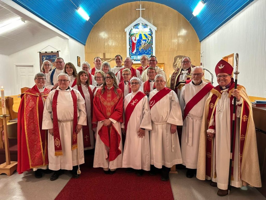 The image shows a group of people standing together. Most of them are wearing vestments, and some are wearing stoles with red accents. In the background, there's a church altar with a stained-glass window depicting a religious scene.