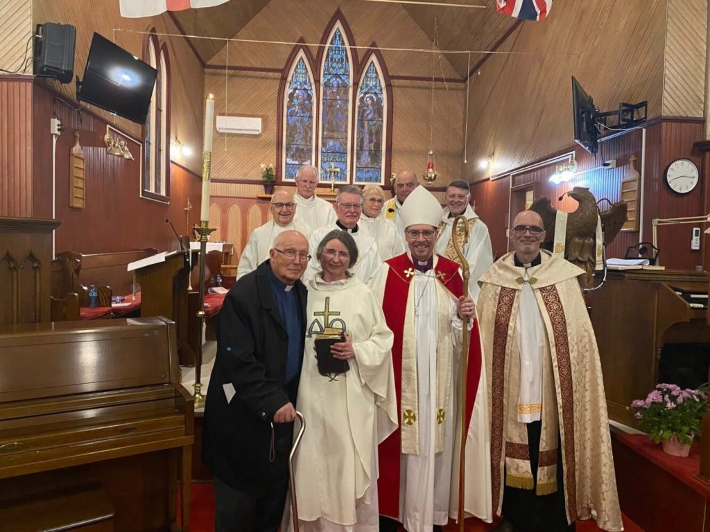 The image shows a group of people standing together. Many of them are wearing white vestments, including one dressed as an Anglican bishop