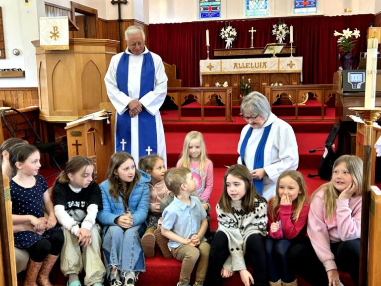 The image shows a group of children and two adults inside a church. The adults are dressed in white robes, and are behind the children, who are sitting on steps