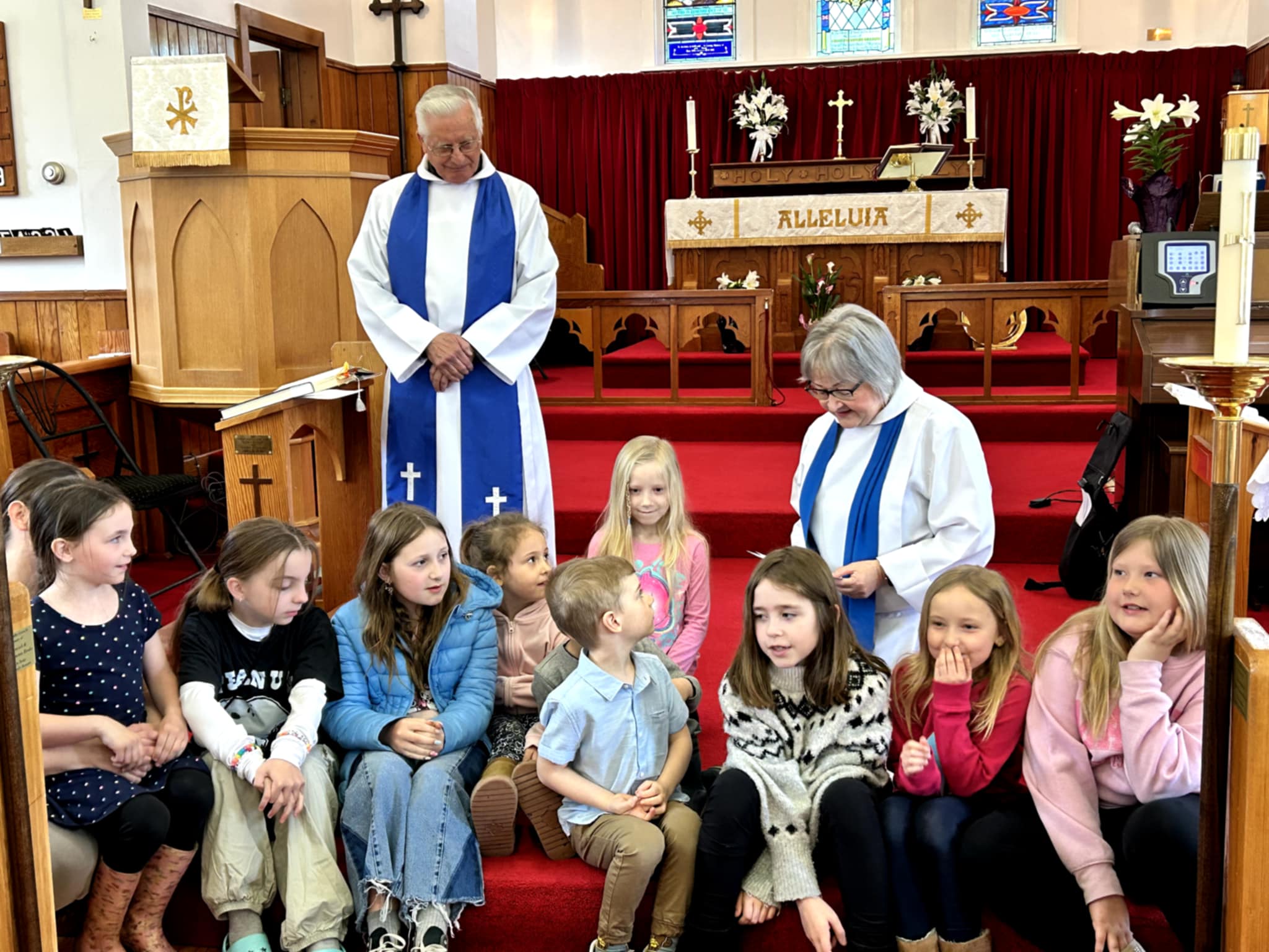 The image shows a group of children and two adults inside a church. The adults are dressed in white robes, and are behind the children, who are sitting on steps