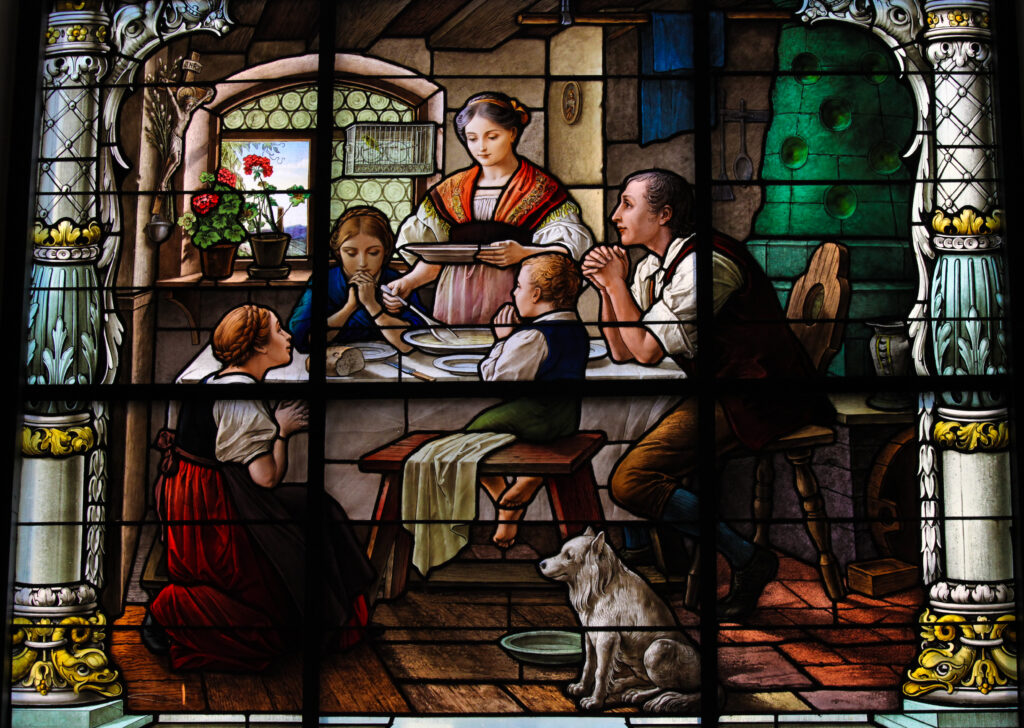 The stained glass window depicts a family gathered around a table, seemingly in prayer before a meal. A woman serves food while others, including children and a man, have their hands clasped. A dog sits patiently by a bowl on the floor. The scene is framed by ornate borders typical of stained glass art.