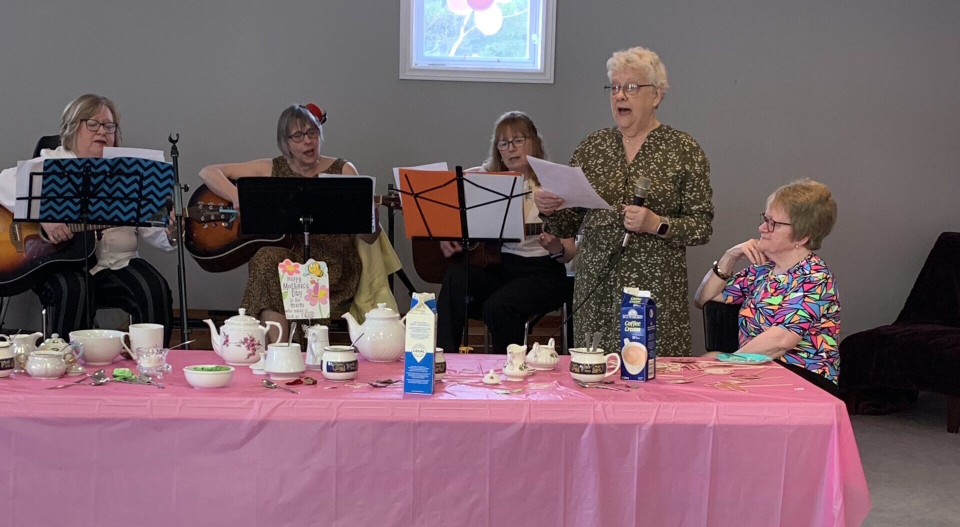 women sitting at a long table with a pink tablecloth on it. one woman reads from a piece of paper.