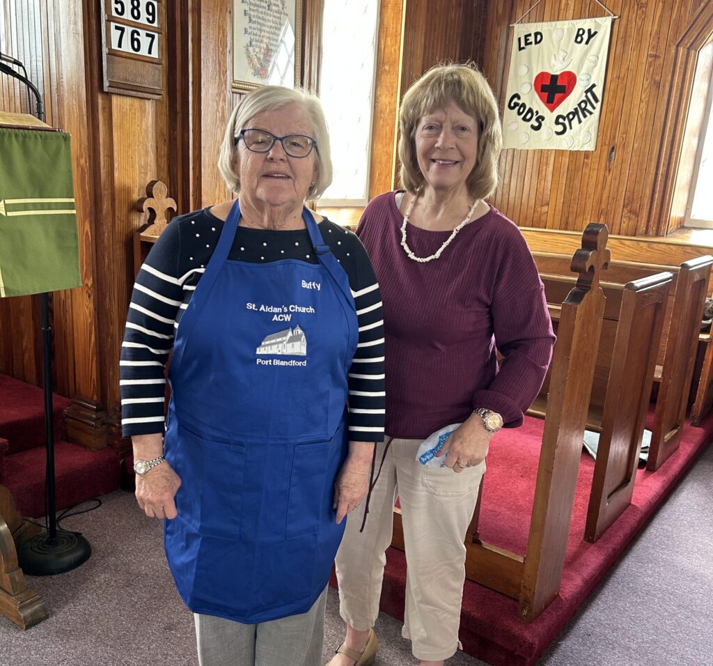two women standing side by side, one wearing a blue apron that says Buffy on it.