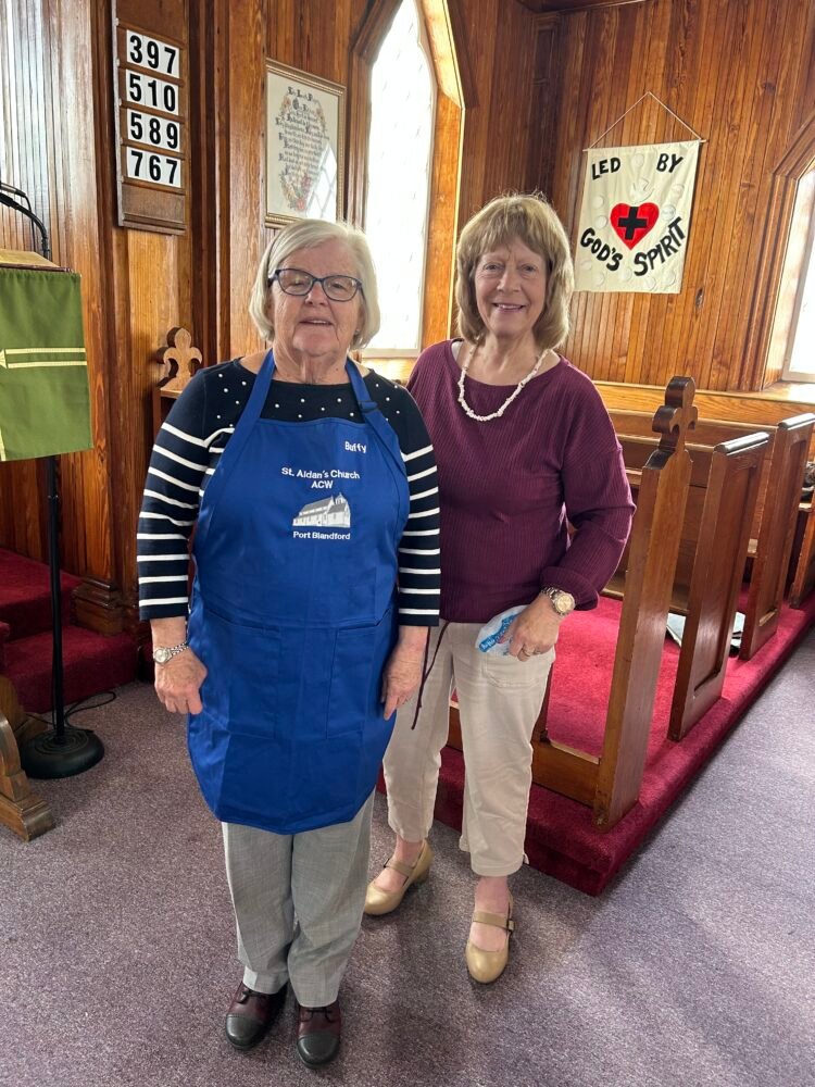 two women standing side by side, one wearing a blue apron that says Buffy on it.