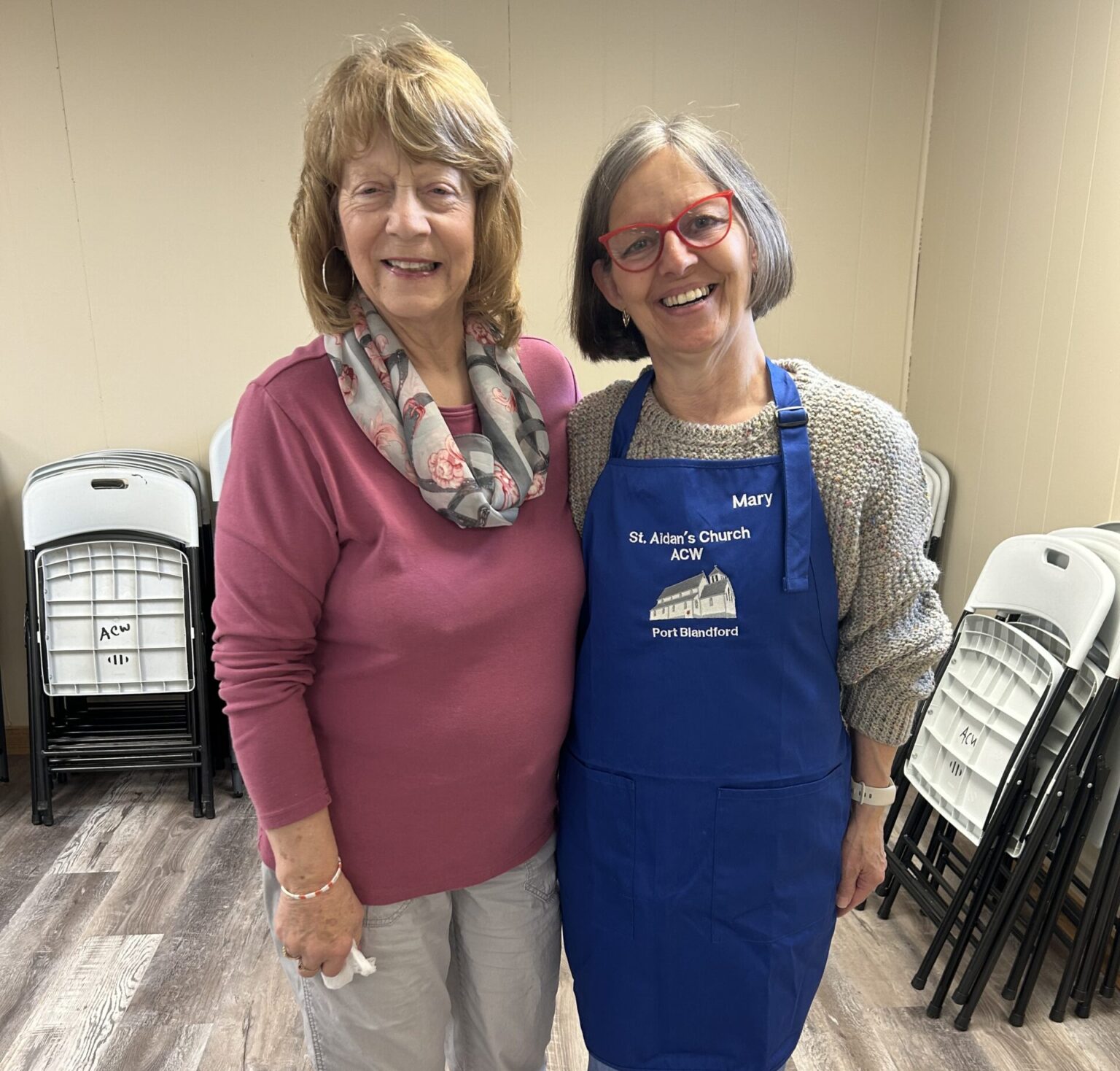 two women standing side by side, one wearing a blue apron that says Mary on it.