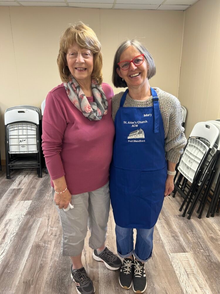 two women standing side by side, one wearing a blue apron that says Mary on it.