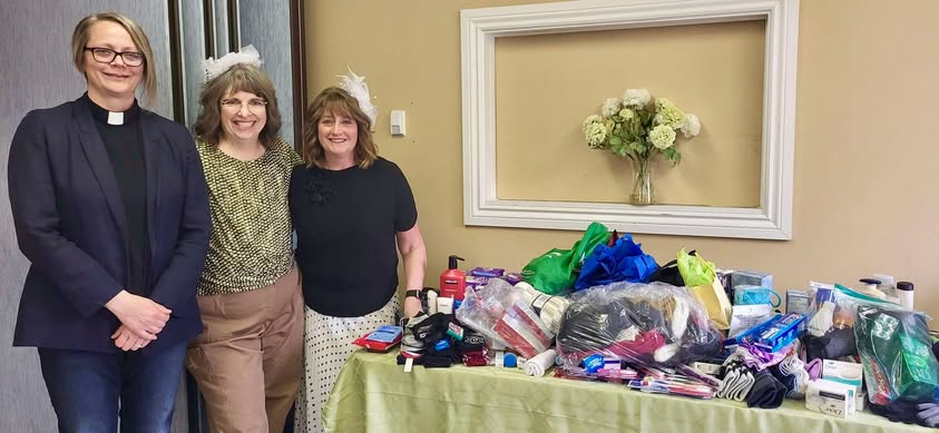 three women next to a table full of socks, hats, mittens, toiletries, feminine hygiene products, and much more