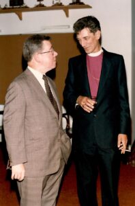 two men, one dressed in a brown suit and one in a black suit, talking to each other