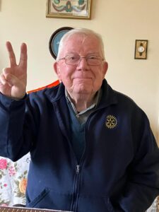 an older man wearing a blue sweater gives the peace sign and smiles