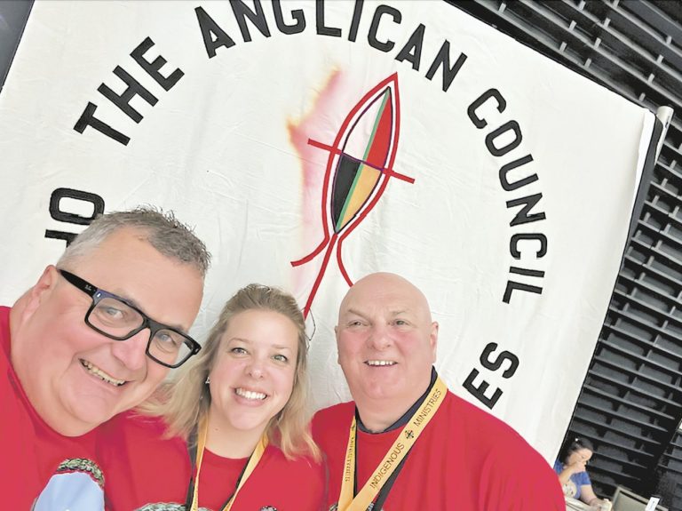 Three people wearing red shirts stand in front of a banner for the Indigenous Church of the Anglican Church of Canada