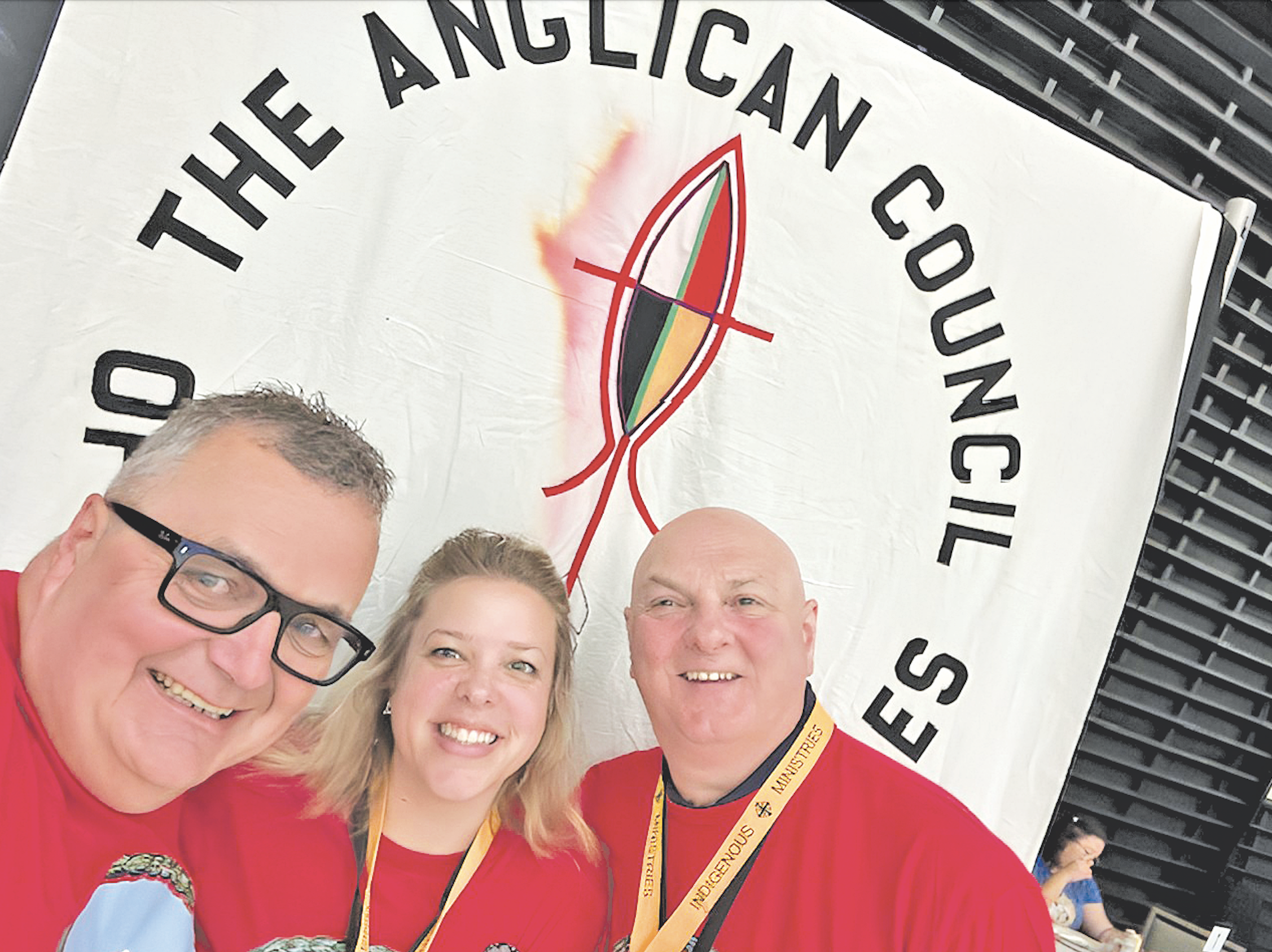 Three people wearing red shirts stand in front of a banner for the Indigenous Church of the Anglican Church of Canada