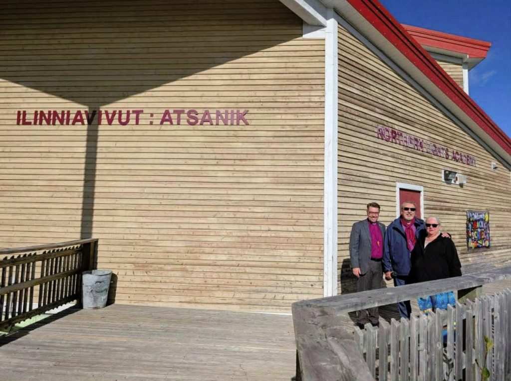 Three people stand outside a wooden school building labeled "ILINNIAVIVUT : ATSANIK" in Nunavik.