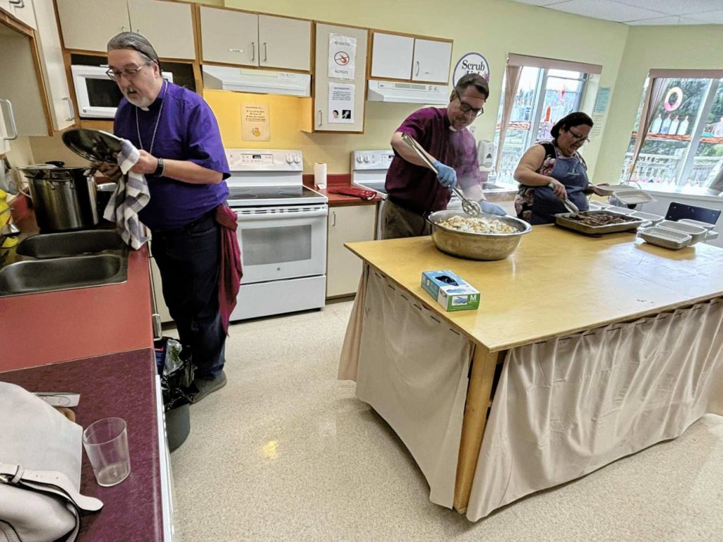 Three people in a kitchen prepare food, stirring and handling trays over a large table.