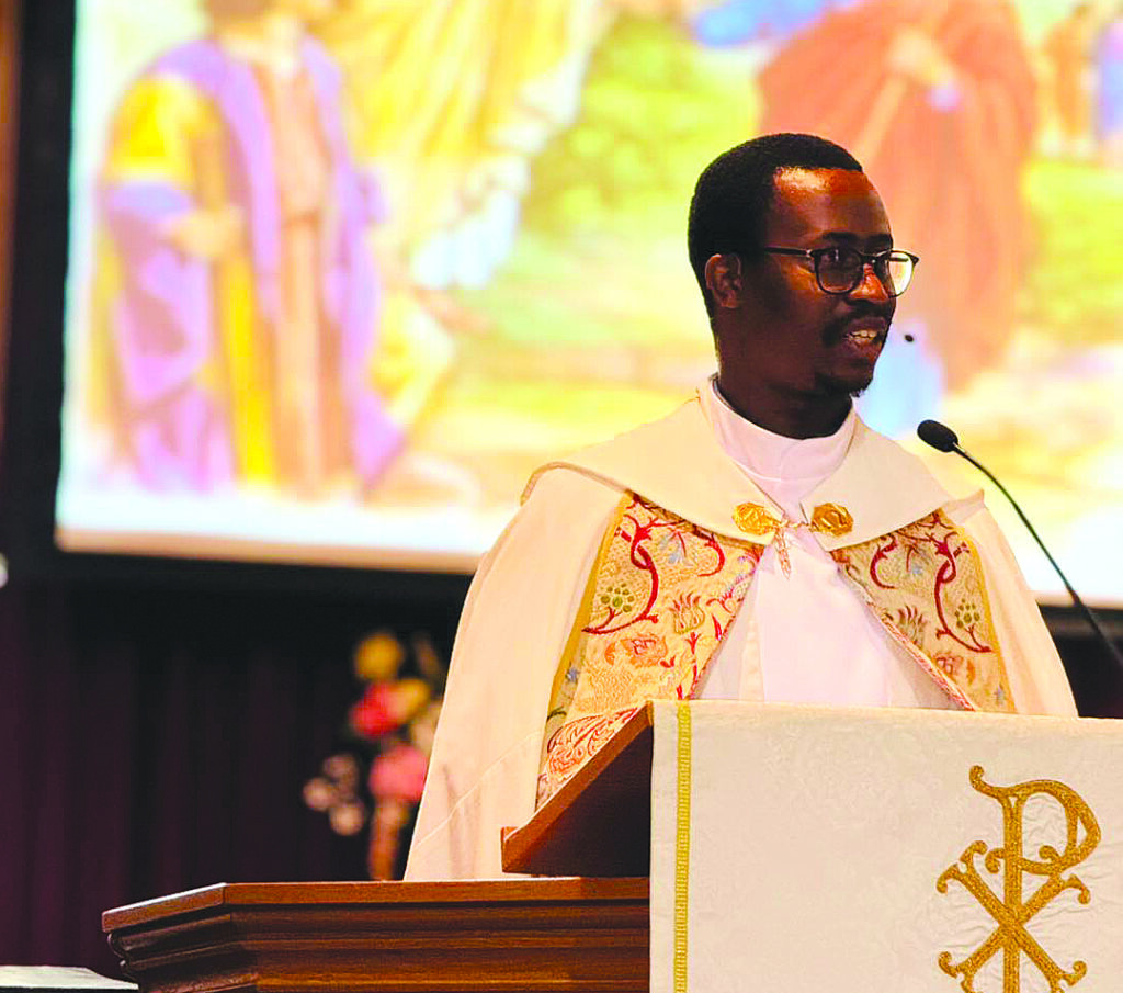 Young man in a white and gold priestly vestment speaking at a pulpit.
