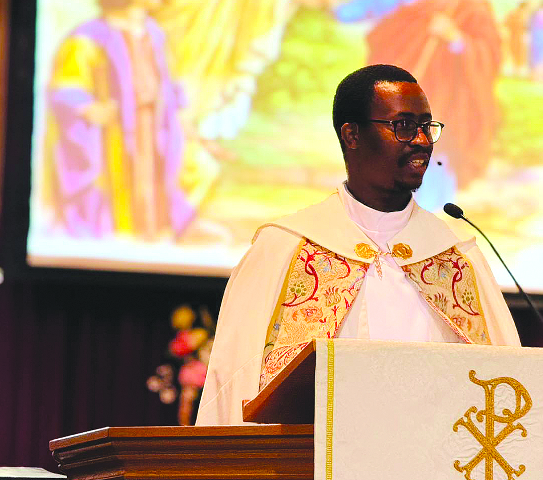 Young man in a white and gold priestly vestment speaking at a pulpit.