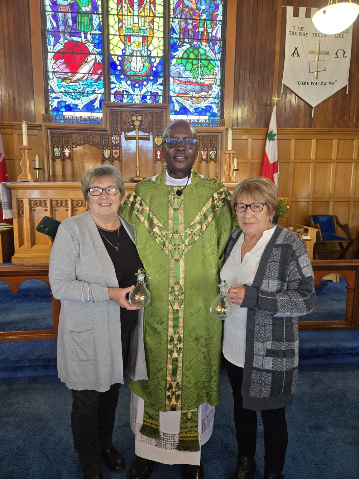 A smiling priest in a green chasuble stands between two women in a church. The women are holding small glass cruets