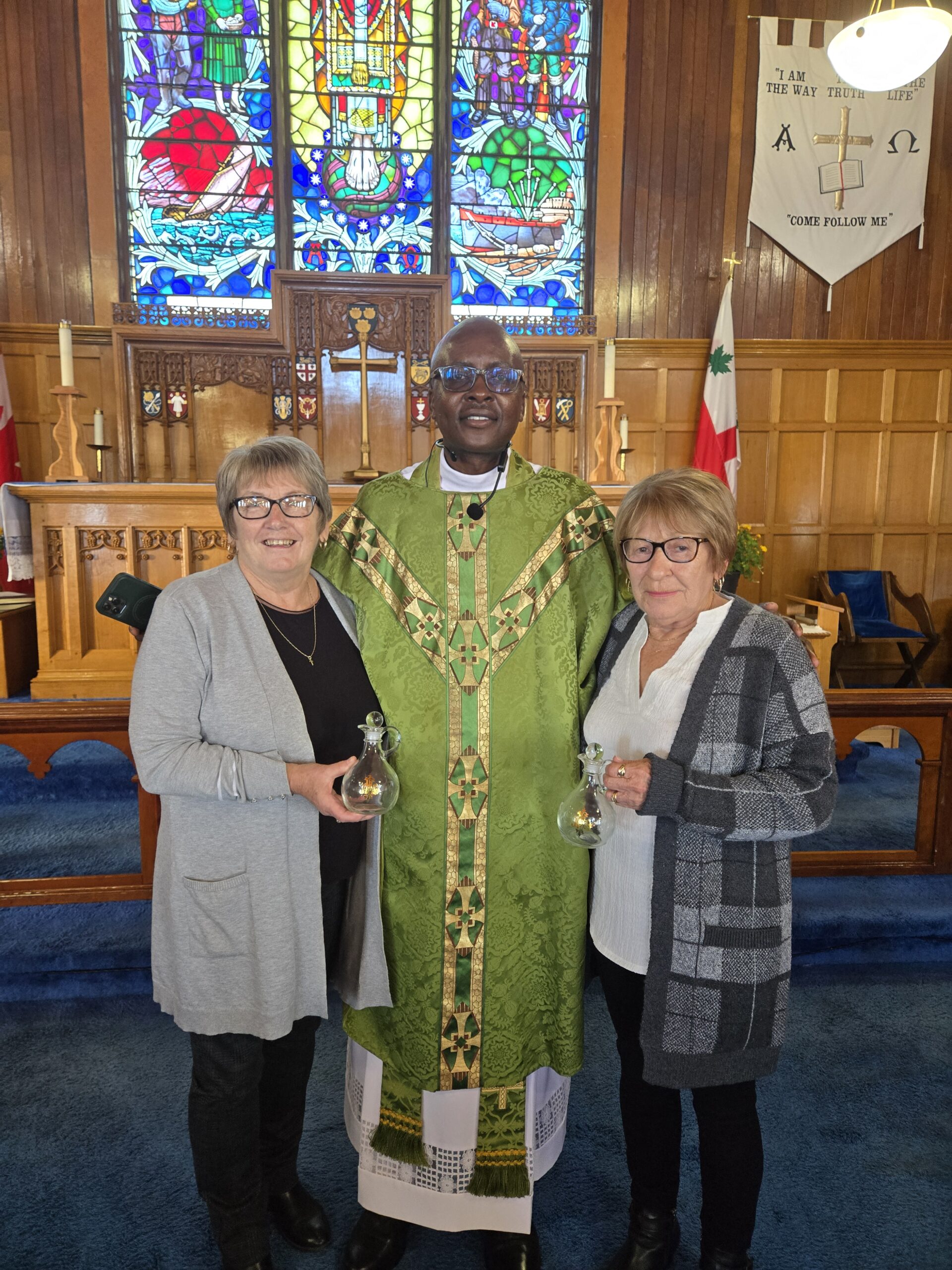 A smiling priest in a green chasuble stands between two women in a church. The women are holding small glass cruets