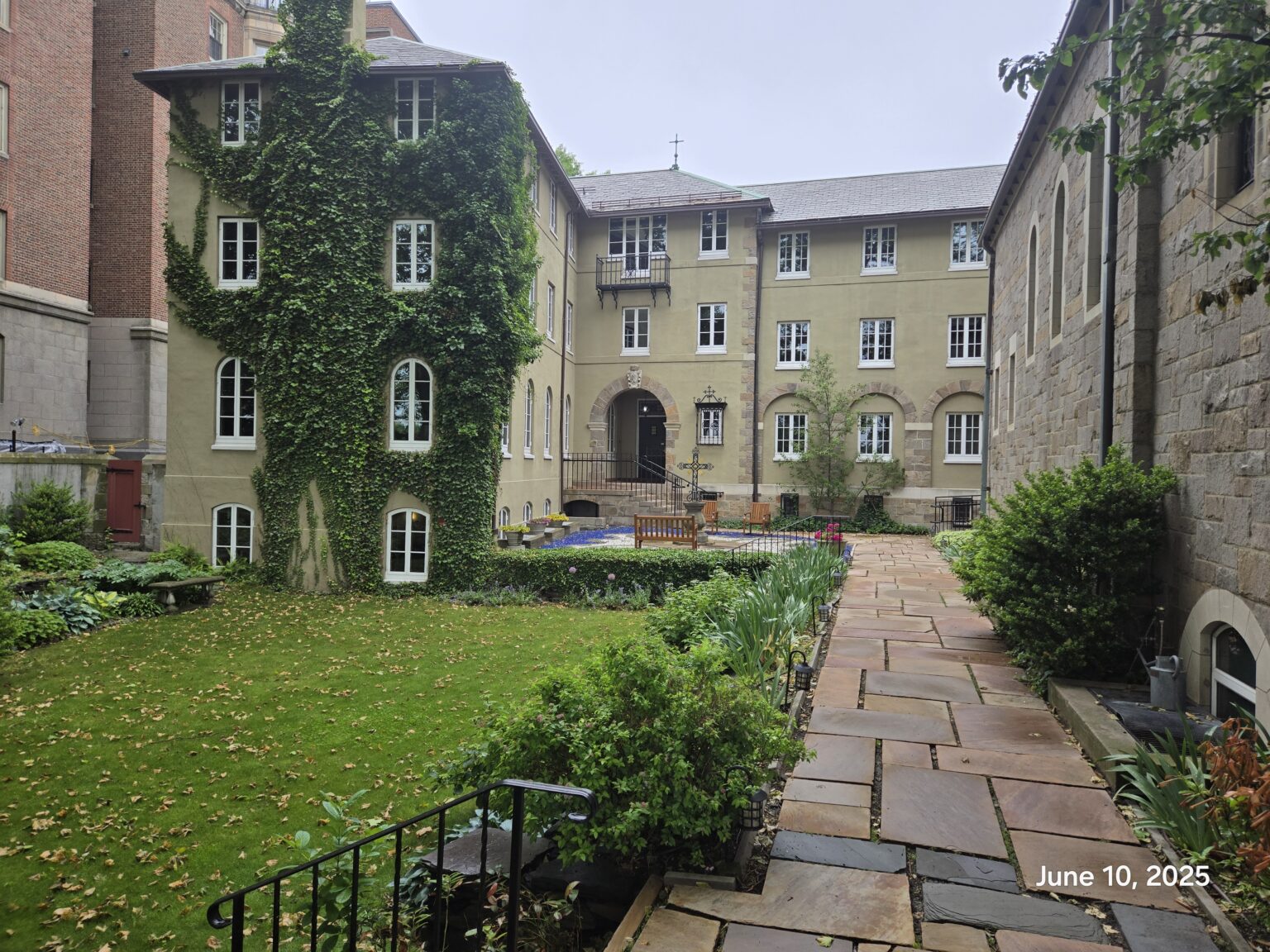 An inner courtyard of a historic building with a stone path leading from the foreground. The building on the left is mostly covered in ivy, and the central building has a covered archway leading into the courtyard.