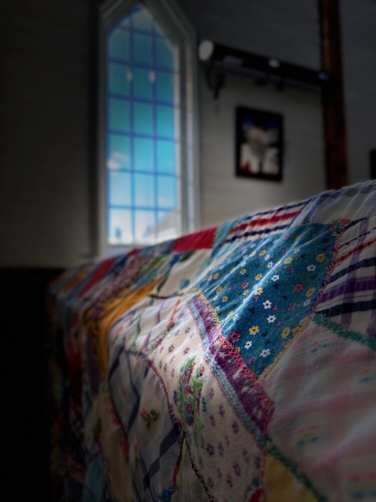 a patchwork quilt draped over a church pew with light coming in a window and falling on it