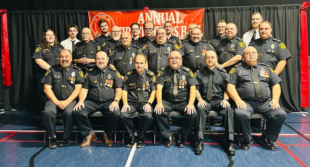 A large group portrait of eighteen volunteer firefighters in dark uniforms, standing and sitting in rows in front of a red banner that reads "Annual Firefighters' Dinner."