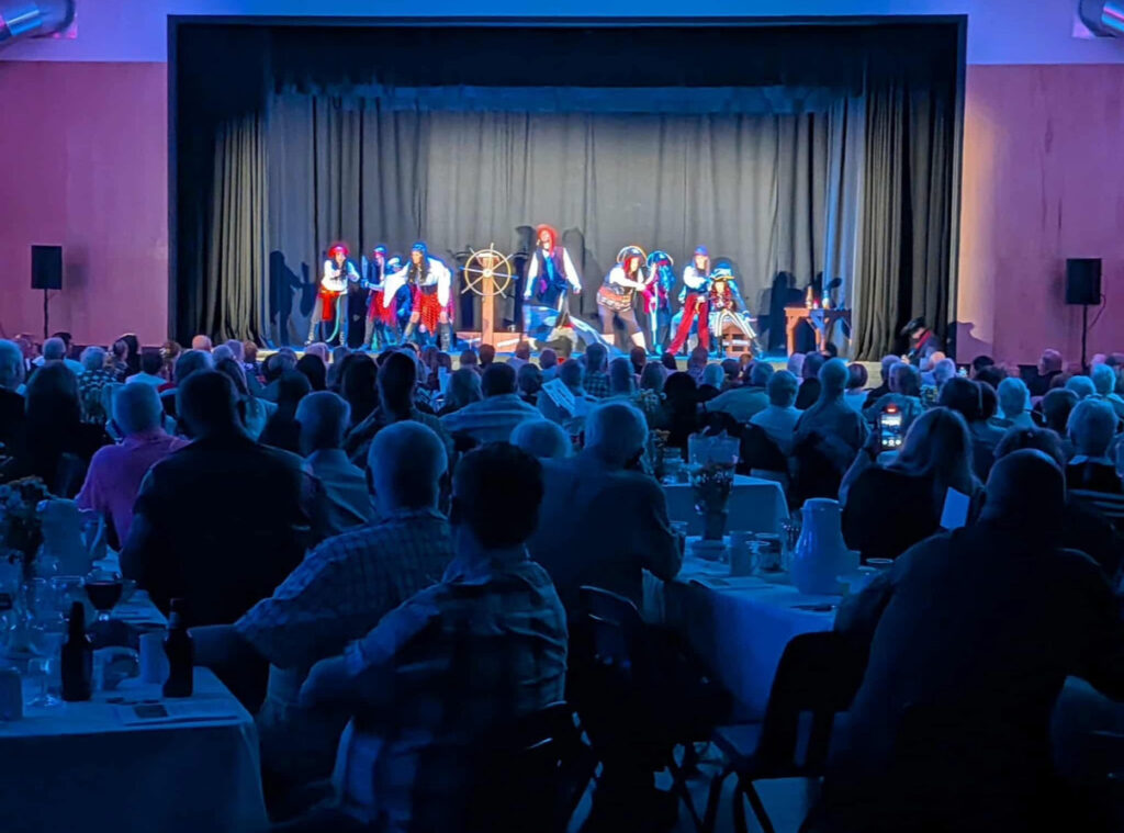 An audience seated at tables in a dimly lit room, looking towards a stage where a group of performers in kilts and colorful costumes are acting out a nautical scene around a ship's wheel.