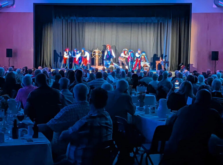 An audience seated at tables in a dimly lit room, looking towards a stage where a group of performers in kilts and colorful costumes are acting out a nautical scene around a ship's wheel.