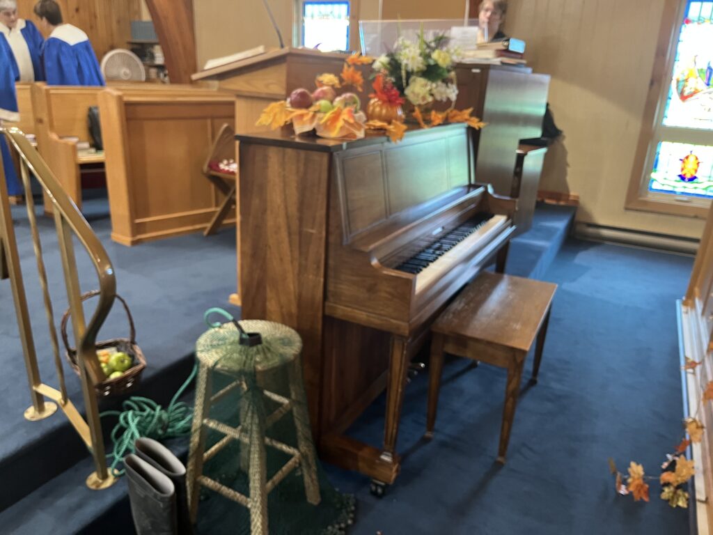 Upright piano in a church, decorated with a harvest display of flowers, gourds, and apples. A person is sitting at the organ console behind it, and a basket of apples and rain boots are on the floor nearby.