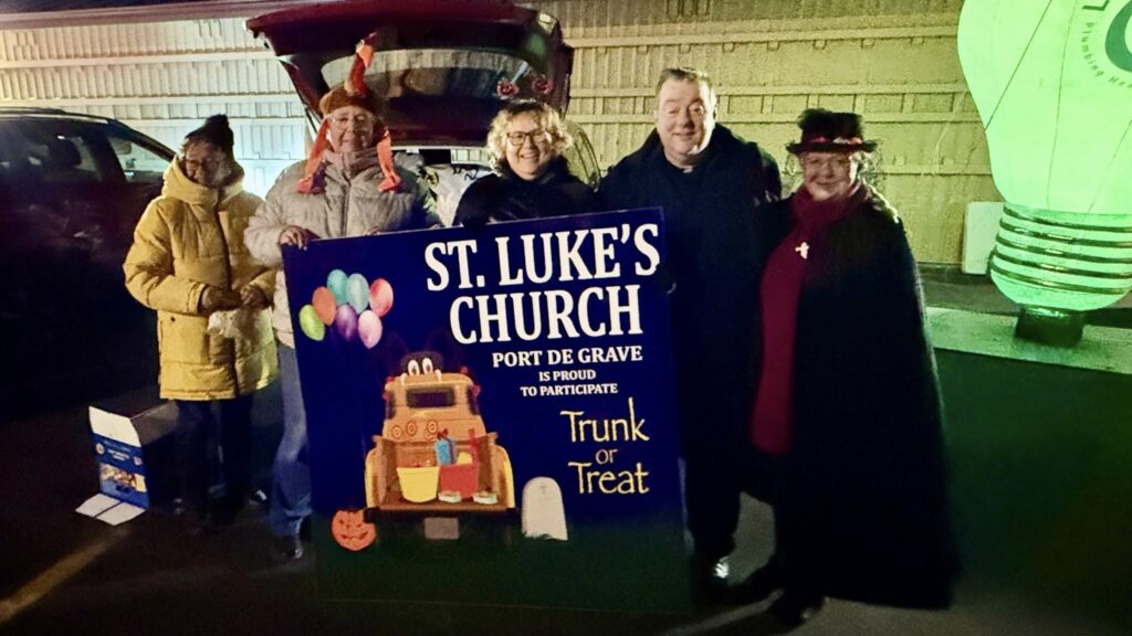 Four adults and a child, some in costume, stand in front of a red SUV holding a large sign for "St. Luke's Church Trunk or Treat." A large, glowing lightbulb prop is visible on the right.