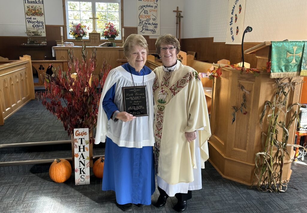 Two women, one wearing a blue and white cassock and the other a white and gold chasuble, stand in a church and smile, holding a commemorative plaque. The church is decorated for a fall harvest with pumpkins, autumn leaves, and a sign that says "GIVE THANKS."