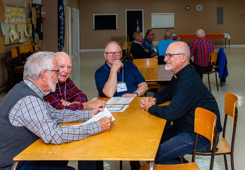 Four men sitting around a wooden table during a small group discussion, with one man on the far left reading from a paper.