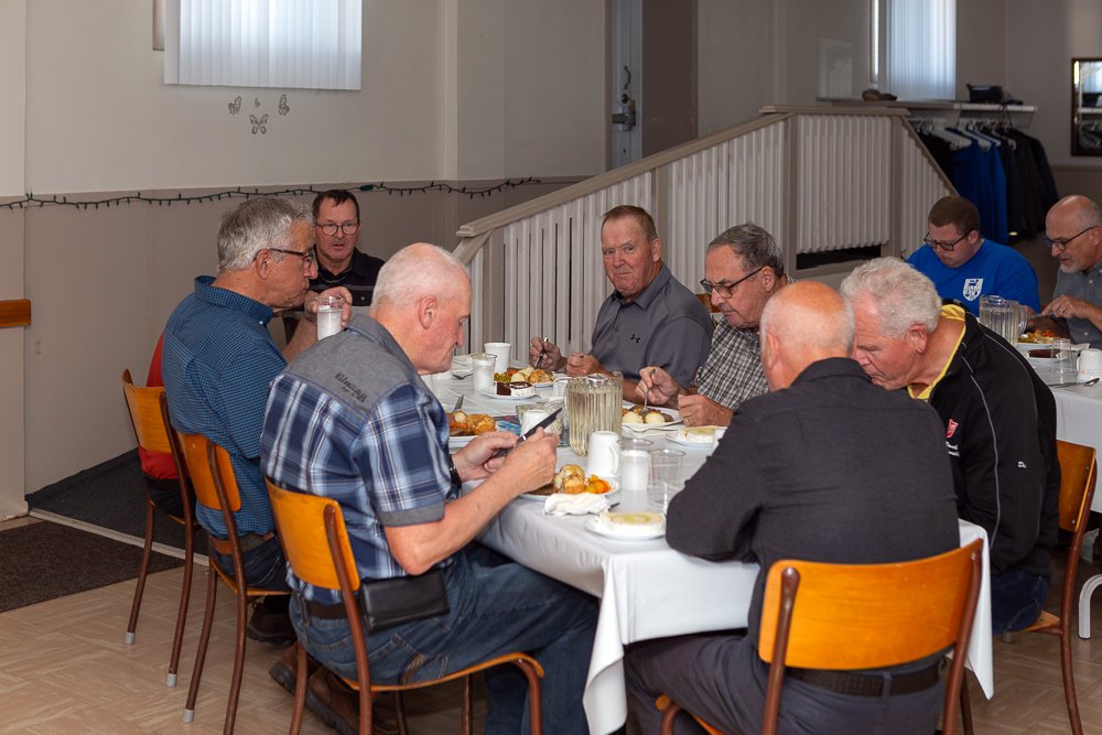 A group of eight men seated around a white-clothed table eating a meal. One man in a plaid shirt is looking at his phone.