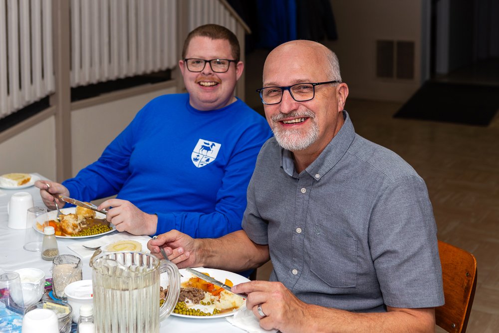Two men seated at a table, smiling at the camera while eating lunch. The man in the foreground has a white beard and glasses, and the man behind him is wearing a bright blue long-sleeved shirt.