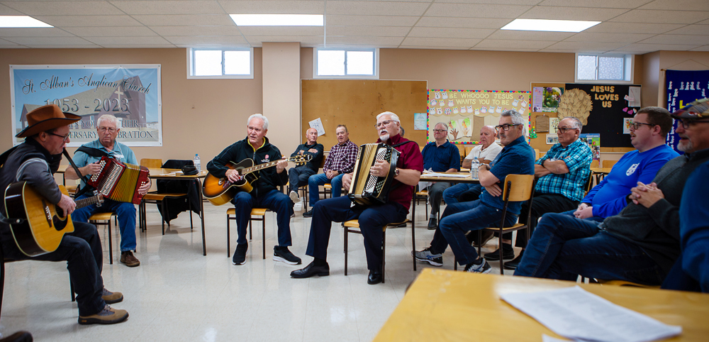 A group of men seated in a semi-circle in a room, playing music. Three men are playing acoustic guitars and one is playing an accordion, while others sit and listen.