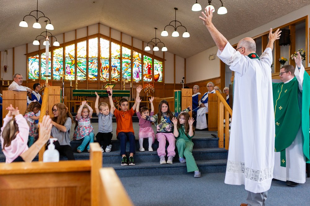 A priest in a white robe and a deacon in a green vestment stand at the front of a church with their hands raised. A group of young children are seated on the steps of the chancel, also with their hands raised in the air. A large stained glass window is visible in the background.