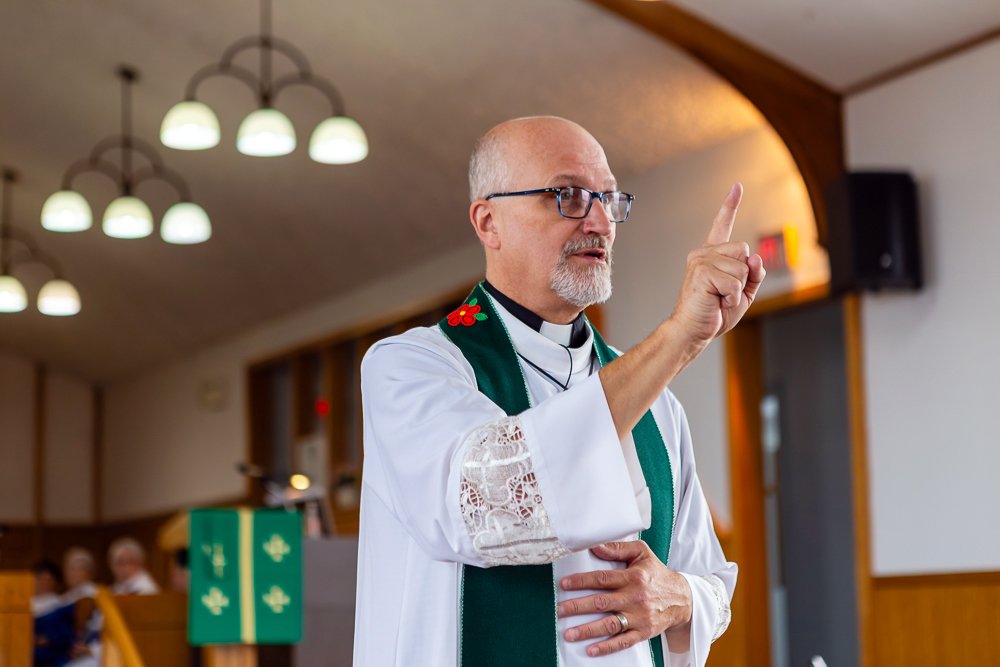 A priest in a white alb with green vestments stands in front of the altar, raising his index finger to make a point while speaking during a church service.