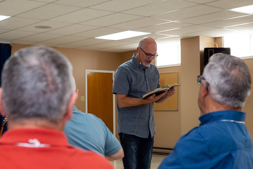 A bald man with a beard and glasses, wearing a short-sleeved blue-gray shirt, stands and reads from a book to an audience whose backs are visible in the foreground.