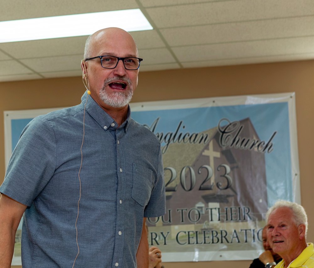 A close-up of a smiling bald man with a beard, wearing glasses and a blue-gray collared shirt, speaking into a lapel microphone. A banner for an "Anglican Church 2023" celebration is visible in the background.