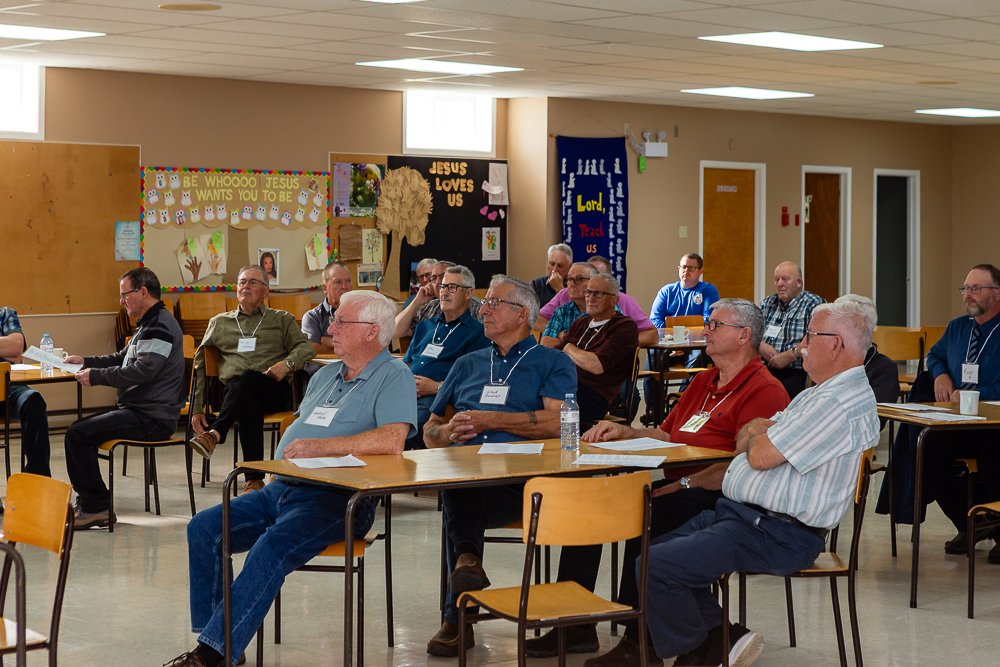 A group of men seated at tables in a large room during a meeting or conference, listening to a speaker off-camera. Religious signage is visible on the back wall.